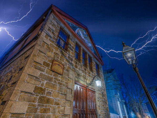 Historic stone building with wooden double doors and barred windows on a downtown street at night, dramatic lightning forks across a deep-blue sky while a glowing vintage lamp post lights the facade.