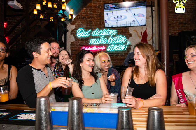 Friends laughing at a neon-lit bar, holding drinks at a wooden counter with exposed brick, a TV showing hockey, and a neon sign reading Bad decisions make better stories!