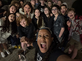 Lively nighttime group selfie of about 15 young people gathered outdoors, crowding around a person in front holding a small metal lantern, everyone smiling, laughing and cheering.