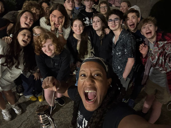 Lively nighttime group selfie of about 15 young people gathered outdoors, crowding around a person in front holding a small metal lantern, everyone smiling, laughing and cheering.