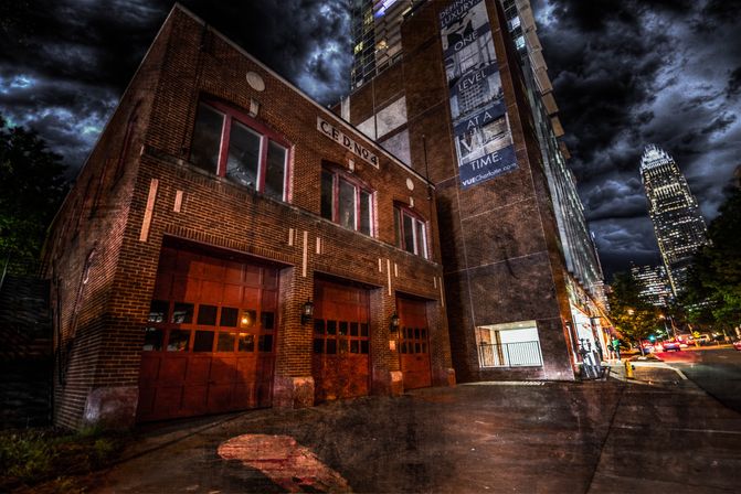 Moody historic brick firehouse with red bay doors at night, dramatic stormy clouds and illuminated downtown high-rise skyline in the background