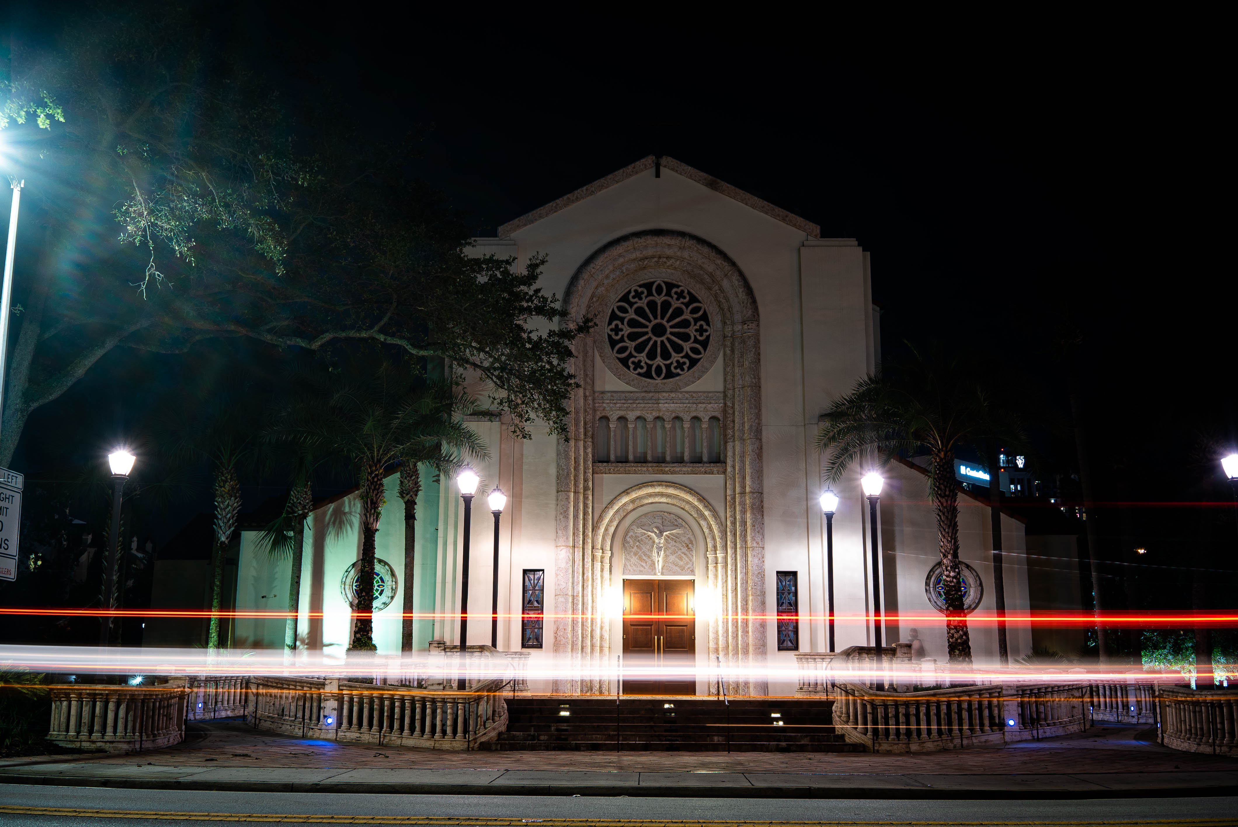 Illuminated historic church façade at night with palm trees and street lamps, long-exposure red and white car light trails streaking across the foreground