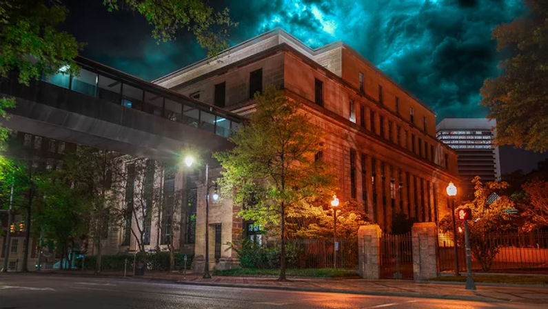 Nighttime urban scene of a historic stone civic building with columns, illuminated street lamps, gated entrance and glass skybridge, trees lining a quiet street under dramatic teal clouds.