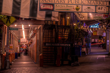 Nighttime scene of a narrow covered market alley with glowing shop signs, string lights, closed metal shutters, cobblestone pavement and a lone pedestrian under warm amber and purple lighting.