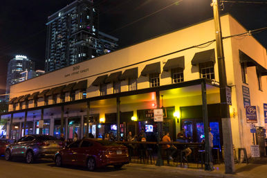 Nighttime downtown nightlife scene: a corner bar with covered outdoor seating and patrons under glowing yellow and purple lights, parked cars on the street and illuminated high-rise towers in the background.