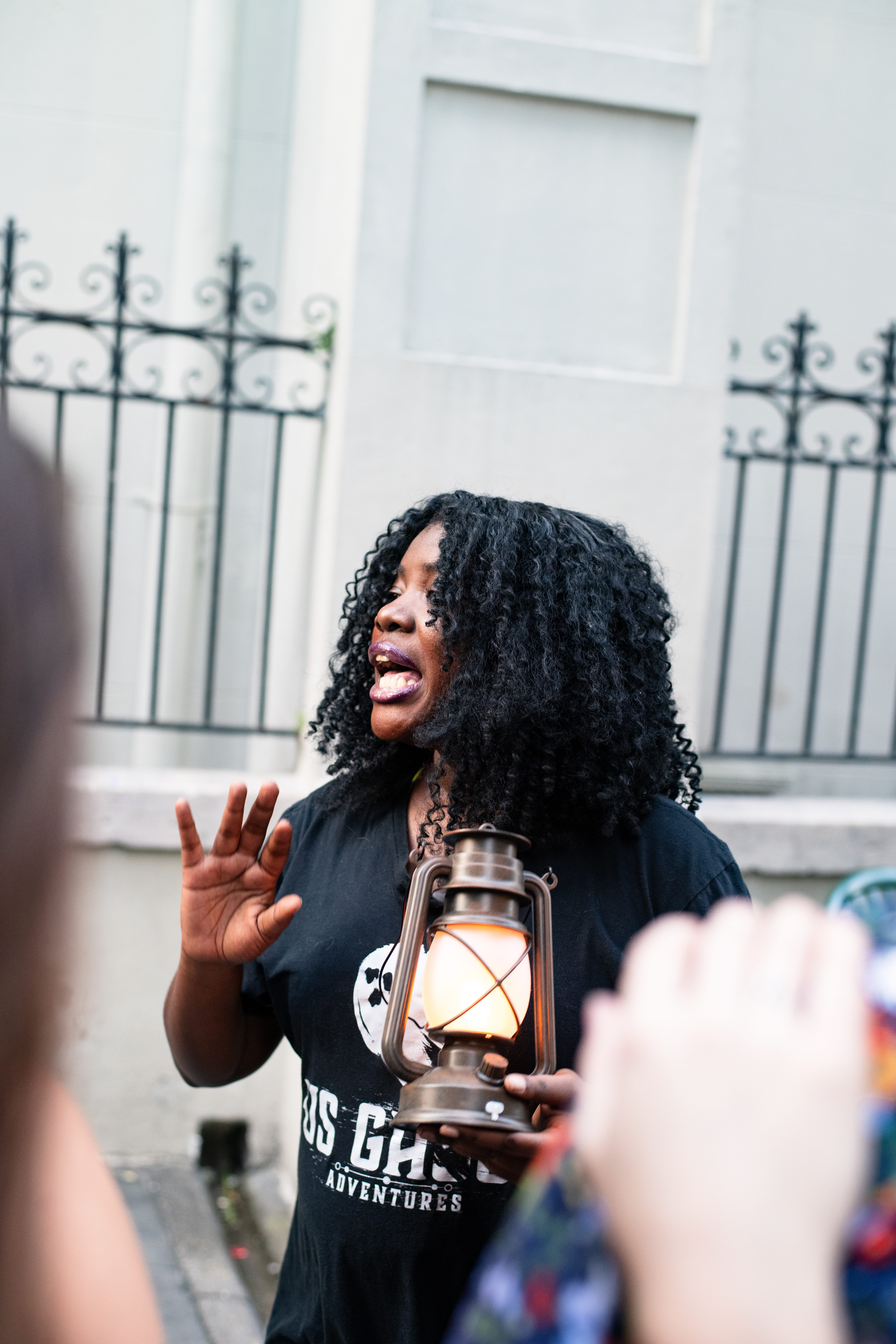 Animated walking tour guide holding a glowing vintage lantern, speaking to a small group on a historic city street in front of a wrought-iron fence.