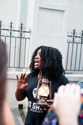 Tour guide holding a glowing vintage lantern, speaking animatedly to a small crowd on a city sidewalk in front of decorative iron fencing and a historic building facade.