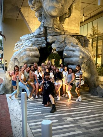 Playful group photo of friends and tourists posing inside and around a giant stone torso sculpture ripping open at the chest to form a tunnel, outdoor urban plaza public-art photo op at dusk.