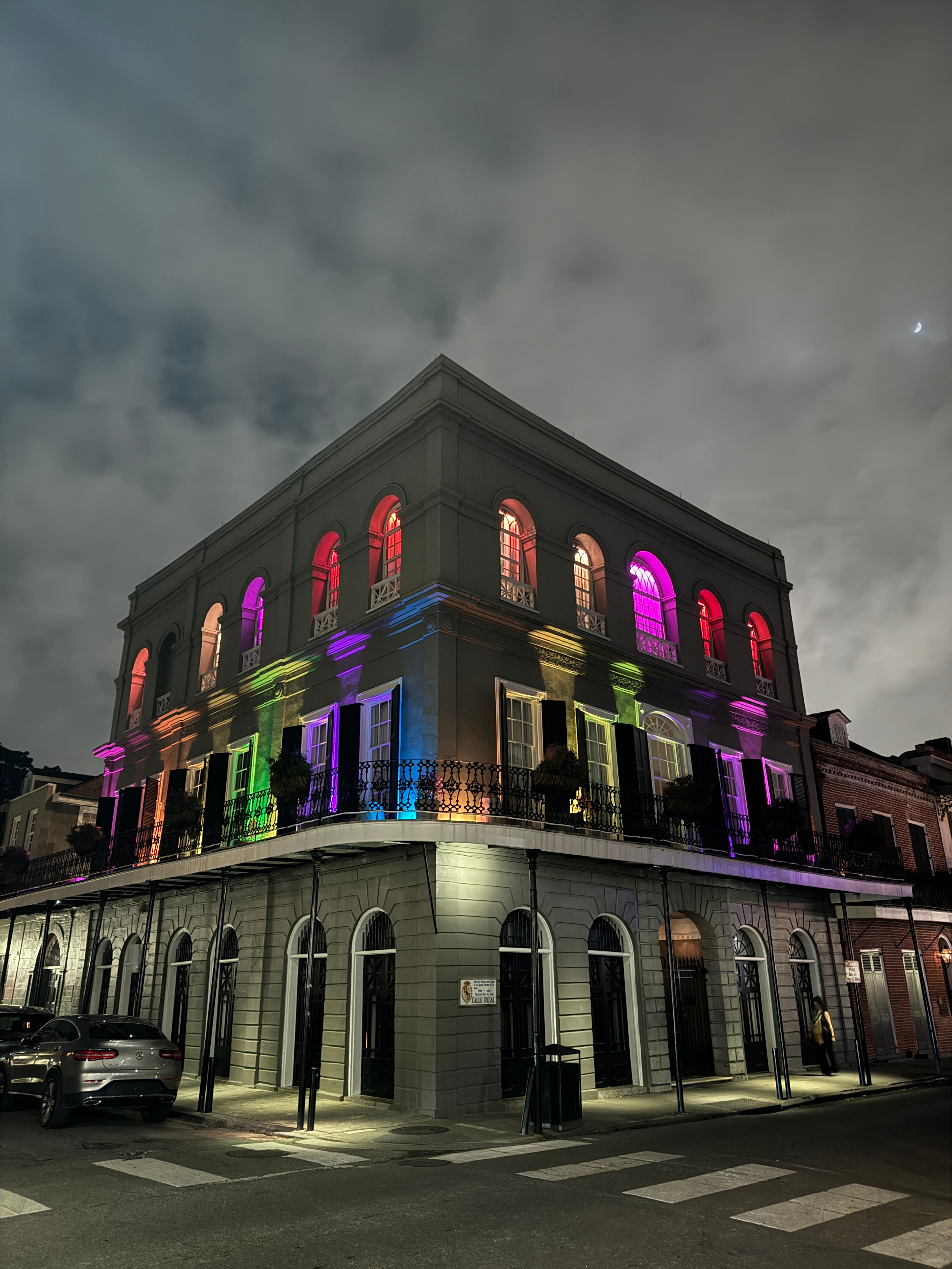 Vibrant night shot of a historic New Orleans French Quarter corner building with arched windows and rainbow-colored lights beneath a cloudy sky and crescent moon.