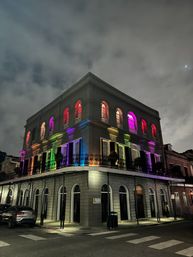 Vibrant night shot of a historic New Orleans French Quarter corner building with arched windows and rainbow-colored lights beneath a cloudy sky and crescent moon.