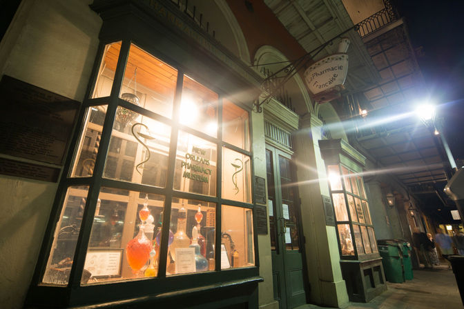 Historic apothecary storefront in New Orleans French Quarter at night, illuminated display of colorful glass apothecary bottles and hanging pharmacy sign.