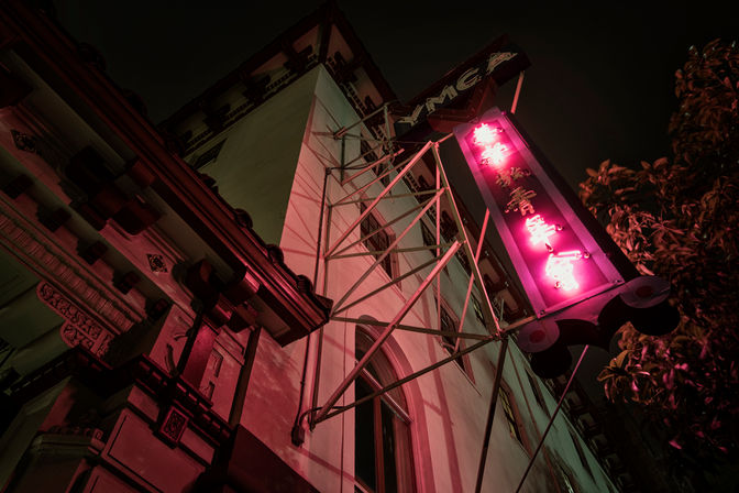 Upward view of a historic urban building at night, pink neon vertical sign with Chinese characters on metal brackets casting magenta light on ornate façade, moody cinematic vibe