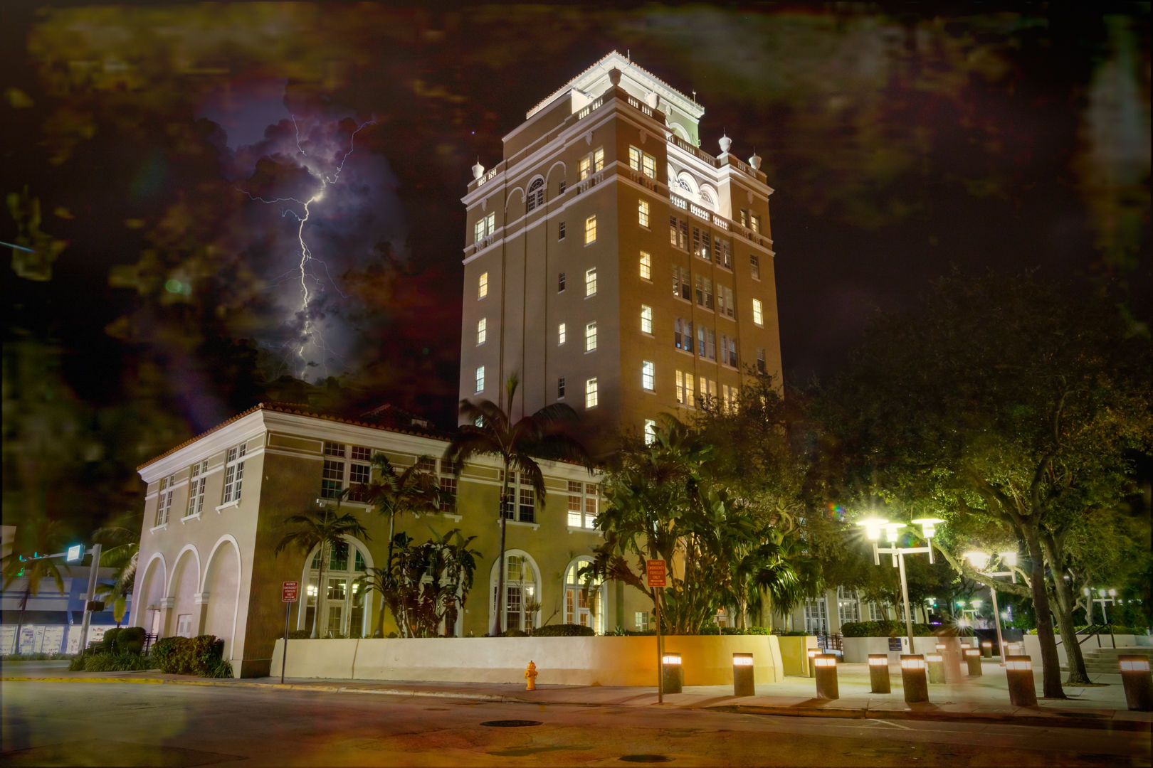 Nighttime view of a historic downtown tower with glowing windows and palm trees, dramatic lightning slicing a stormy sky above a lit plaza and streetlights.