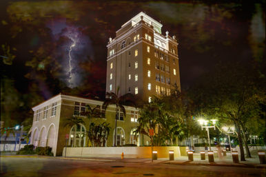 Nighttime view of a historic downtown tower with glowing windows and palm trees, dramatic lightning slicing a stormy sky above a lit plaza and streetlights.