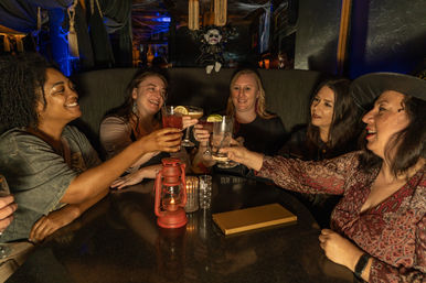 Five friends toasting cocktails in a dimly lit bar booth at night, lantern and shot glass on the table, with Halloween doll and spiderweb decorations in the background.