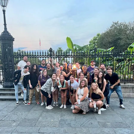 Group of about two dozen friends smiling and posing at dusk in an urban plaza in front of a decorative wrought-iron fence and tropical banana plants, many holding drinks — lively outdoor group photo.