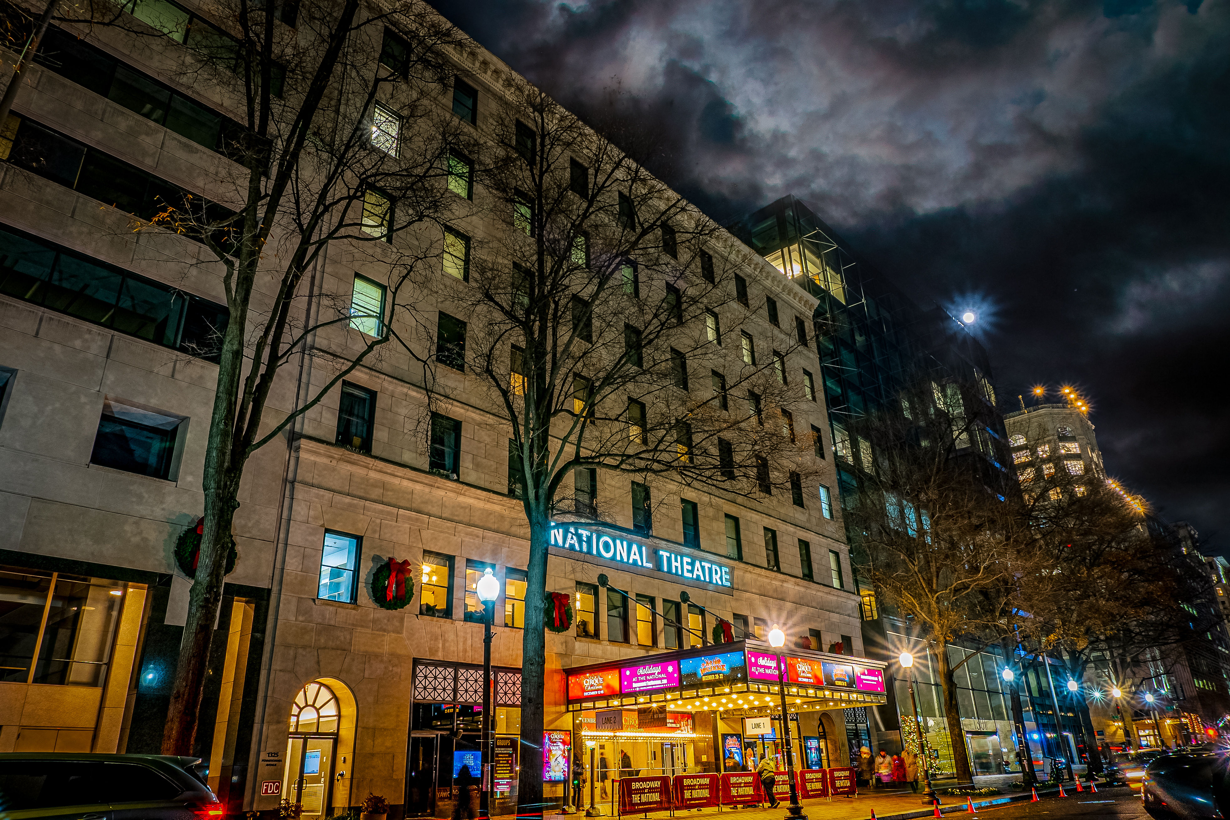 Urban stone theater with glowing marquee and holiday wreaths, leafless trees and cars along a busy city street under a dramatic cloudy night sky
