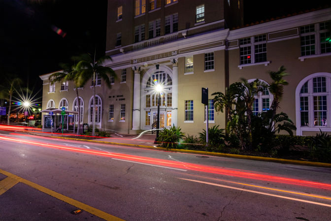Historic city hall at night with arched windows and palm trees; long-exposure red and white light trails streak across the downtown street.
