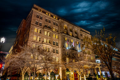Historic stone hotel on a downtown city street at night, warm glowing windows and flags on the façade under a dramatic stormy sky