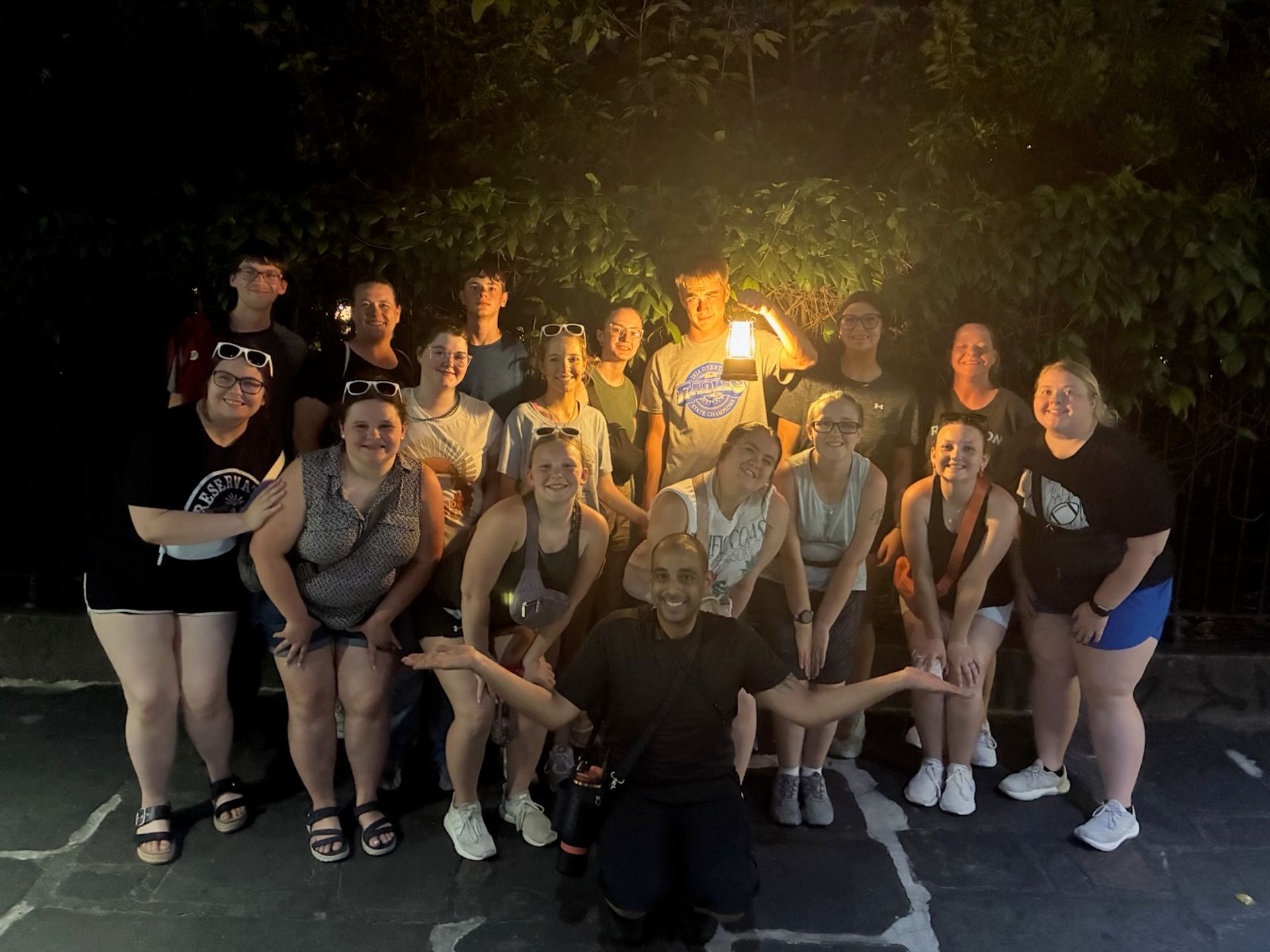 Lantern-lit group photo at night—smiling people posing on stone pavement beneath an ivy-covered wall, warm light casting soft shadows.