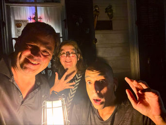 Playful nighttime porch selfie of three adults at a residential front door, faces lit by a warm lantern as they smile and show surprised expressions.