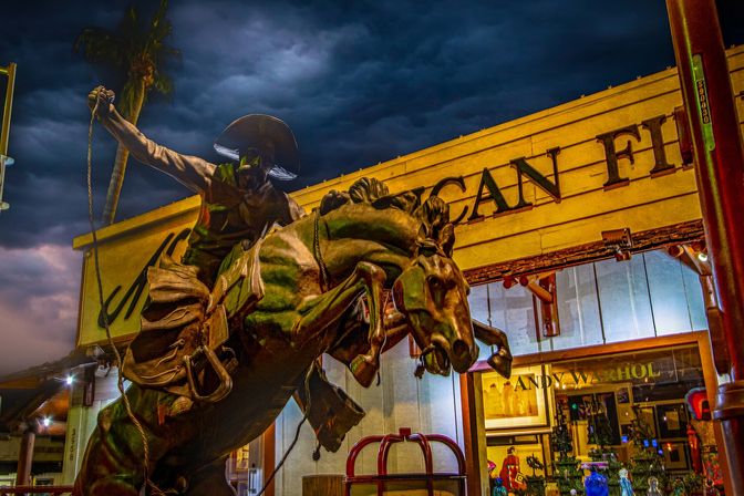 Bronze cowboy statue on a rearing horse outside a brightly lit storefront at night, dramatic stormy sky and palm tree in the background on a downtown tourist shopping street