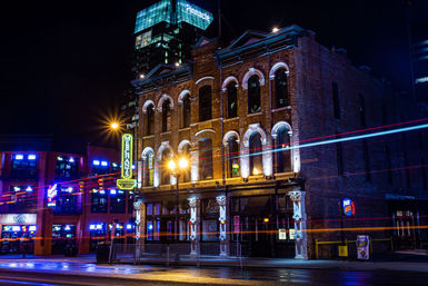 Historic three-story brick building lit up at night in a downtown entertainment district, neon signs and bar windows glowing, streaking car light trails and wet street reflections with a modern glass skyscraper towering behind.