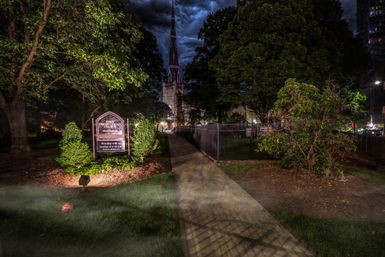 Moody nighttime churchyard in a downtown setting: lit sign and manicured shrubs beside a concrete path leading to a tall gothic steeple framed by trees under dramatic stormy clouds.