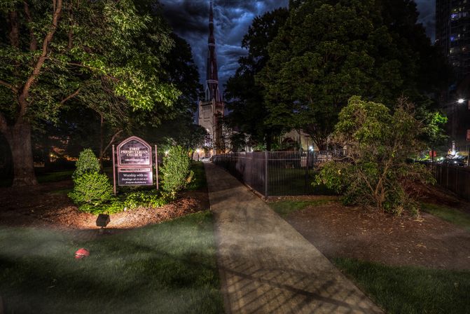 Moody nighttime churchyard in a downtown setting: lit sign and manicured shrubs beside a concrete path leading to a tall gothic steeple framed by trees under dramatic stormy clouds.