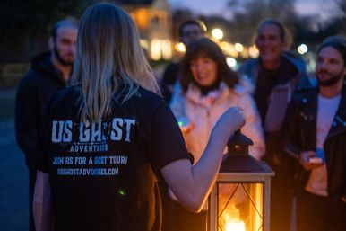 Guide holding a glowing lantern leads a small group on a spooky evening ghost tour outdoors along a dimly lit street
