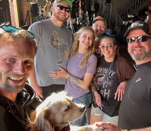 Cheerful group of six adults and a golden retriever taking a selfie inside an indoor live music venue, stage and drum kit visible in the background.