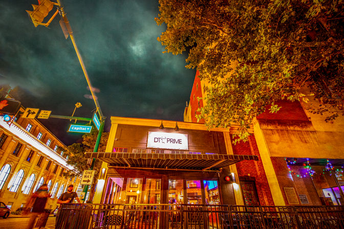 Nighttime downtown street scene on Capitol Street featuring a glowing restaurant storefront with outdoor patio seating, dramatic cloudy sky, and illuminated historic buildings.