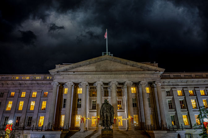 Nighttime view of a neoclassical government building with illuminated columns and windows, a statue on the front steps, an American flag above, stormy clouds overhead and festive holiday garlands and lights