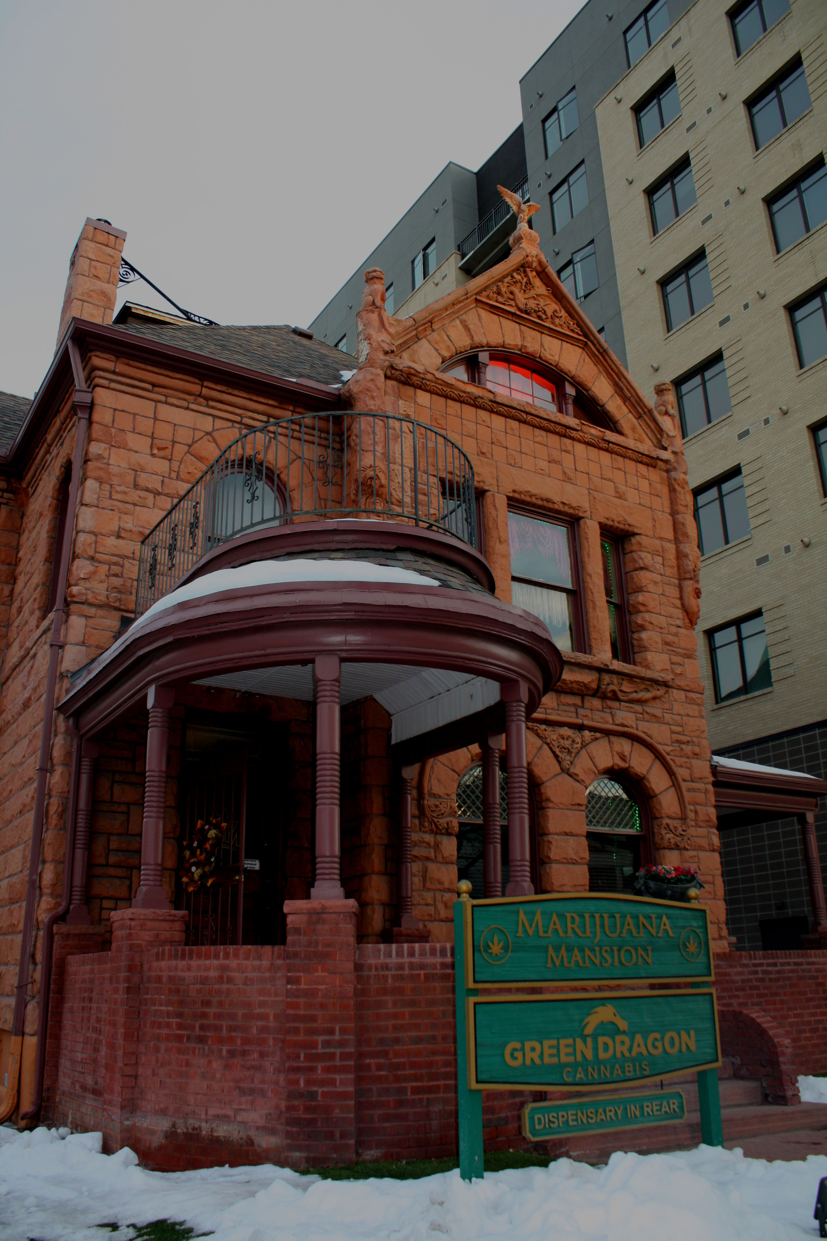 Red sandstone Victorian mansion with rounded porch and wrought-iron balcony in an urban setting, green wooden sign marking a cannabis dispensary out front and light snow on the ground