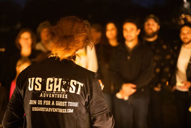 Spooky night ghost tour guide seen from behind addressing a small outdoor group under warm orange light, blurred listeners in the background.