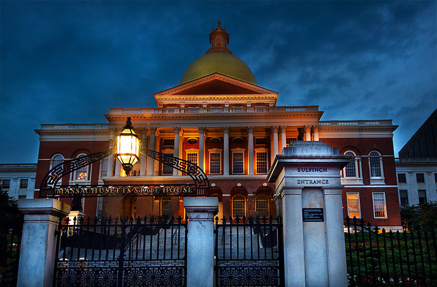 Boston state capitol with a glowing gold dome at dusk, classical columned façade lit warmly behind an ornate iron gate and lantern