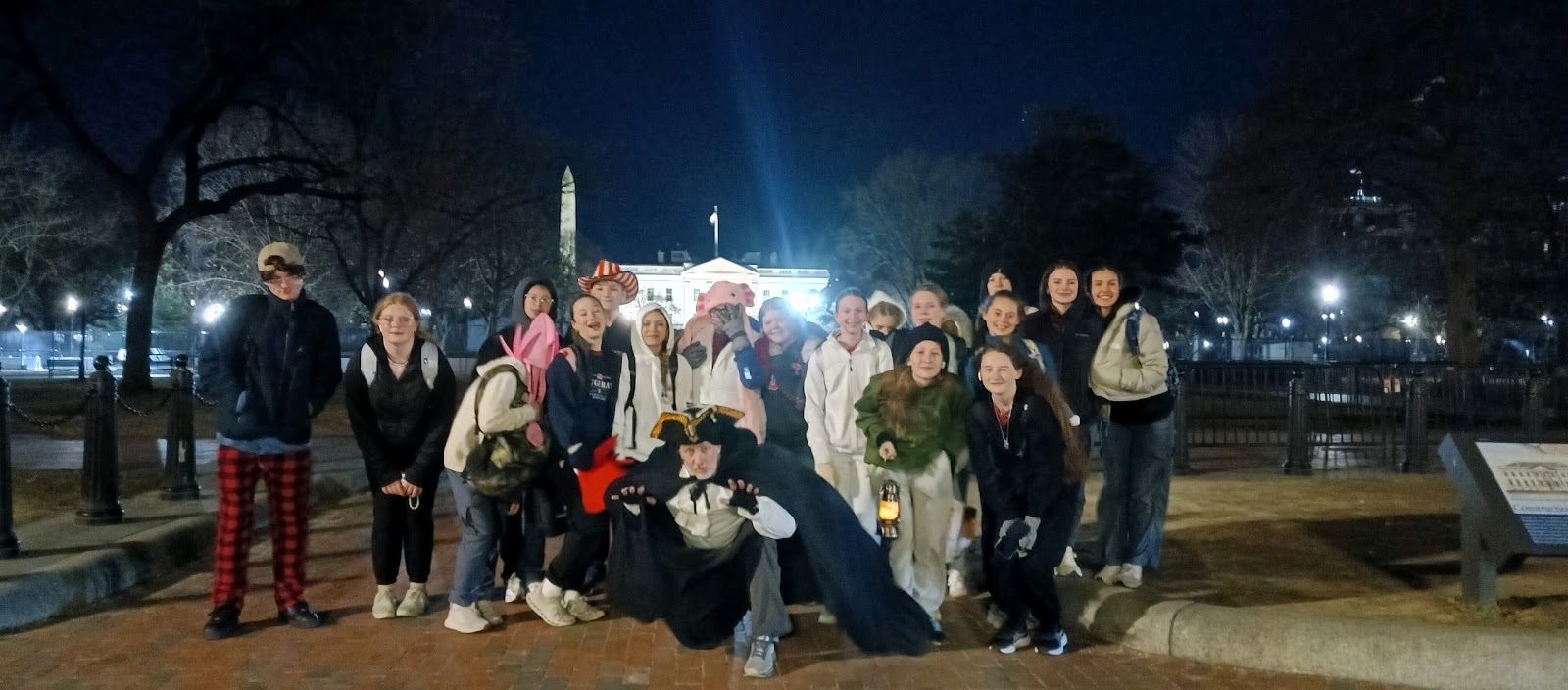 Smiling group of young people on a nighttime lantern tour in front of the White House, Washington Monument lit in the background, with a costumed guide in a tricorne hat crouching and holding a lantern — Washington, D.C.