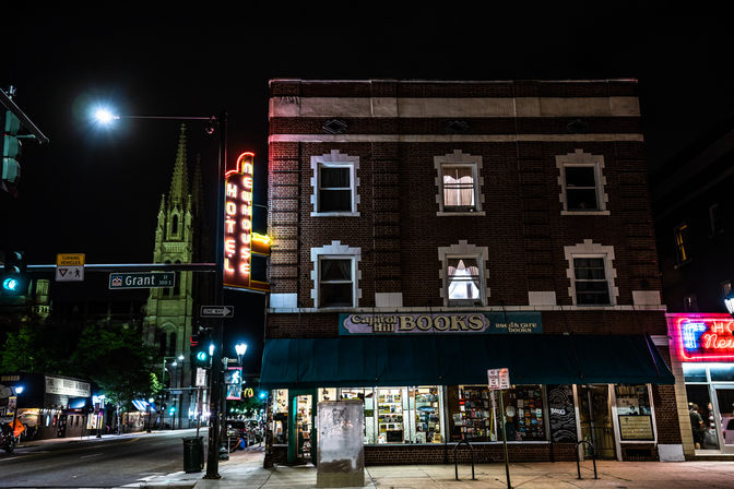 Night city corner with a brick three-story building housing a cozy independent bookstore under a green awning, glowing vertical neon hotel sign on the side, illuminated shop windows and a gothic church steeple in the background.