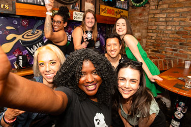 Smiling group selfie of seven friends in a lively brick‑walled bar with a colorful mural and drinks — fun nightlife scene and celebration.
