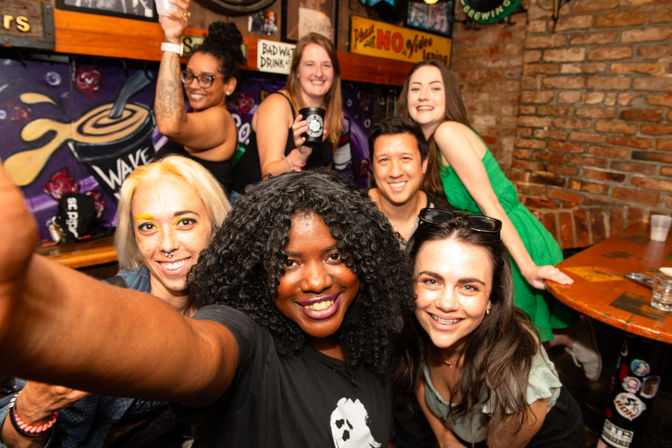 Smiling group selfie of seven friends in a lively brick‑walled bar with a colorful mural and drinks — fun nightlife scene and celebration.