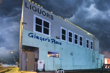 Two-story corner liquor store with pale-blue exterior and string lights, small entrance with illuminated open sign, dramatic storm clouds overhead and an empty night street.