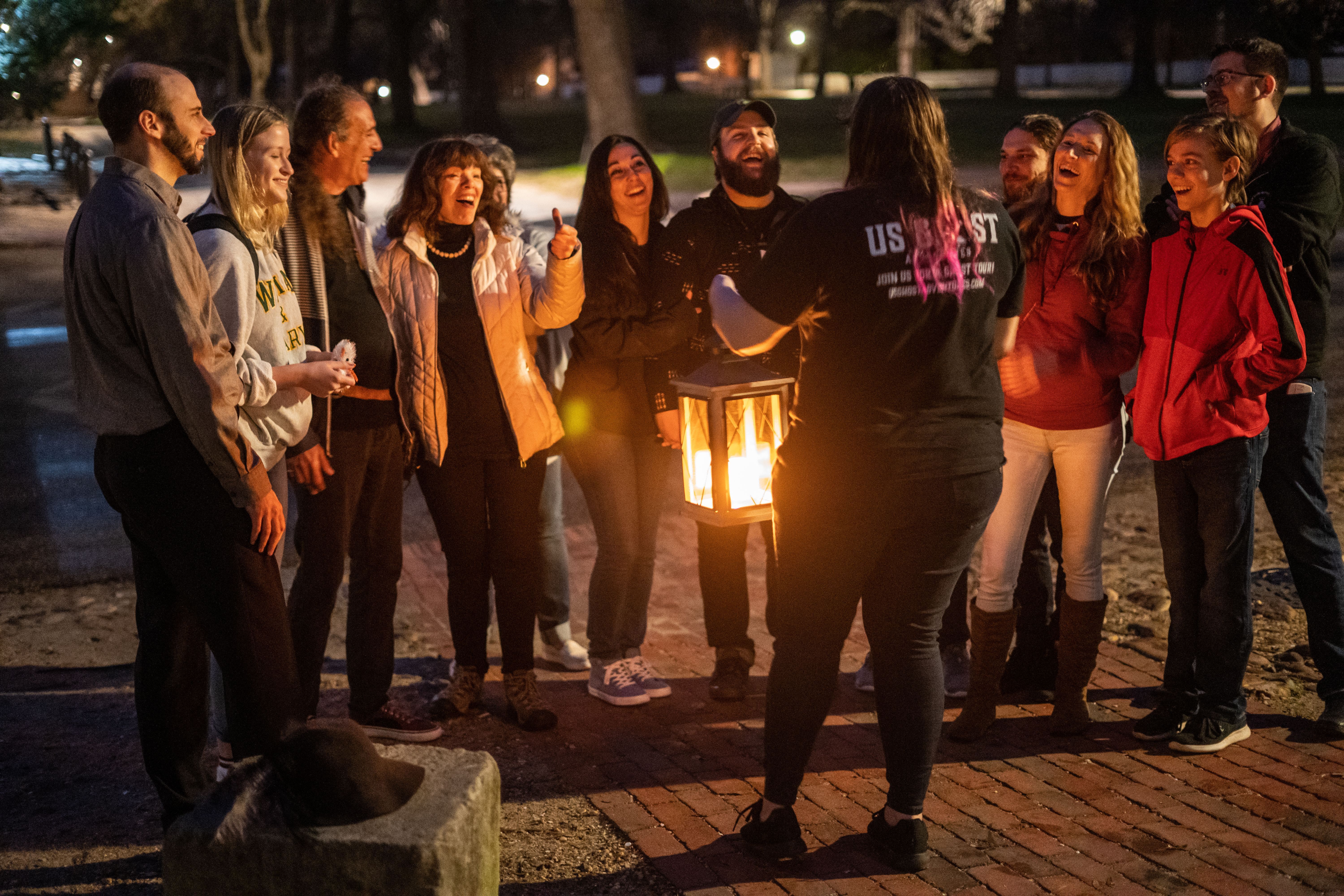 Group of people laughing and listening as a guide holds a glowing lantern on a brick path in a dimly lit park at night.