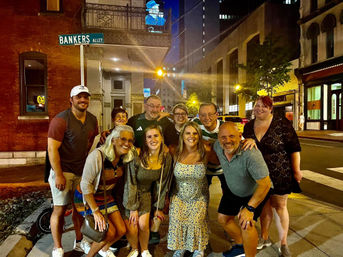 Cheerful group of friends posing on a downtown sidewalk at night under a 'Bankers Alley' street sign, surrounded by city lights, brick buildings, and nightlife