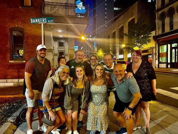 Cheerful group of friends posing on a downtown sidewalk at night under a 'Bankers Alley' street sign, surrounded by city lights, brick buildings, and nightlife