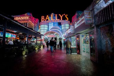 Neon-lit Las Vegas Strip at night with pedestrians walking toward a glowing casino entrance, flanked by outdoor bars, restaurants, and colorful signage.