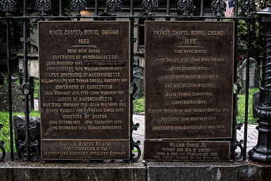Weathered bronze memorial plaques on an ornate wrought-iron gate at a historic 1630 burial ground, listing early colonial-era burials with old headstones and grass visible behind.