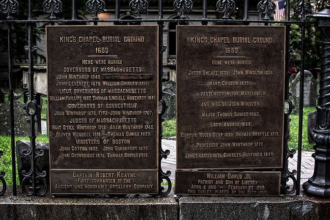 Weathered bronze memorial plaques on an ornate wrought-iron gate at a historic 1630 burial ground, listing early colonial-era burials with old headstones and grass visible behind.