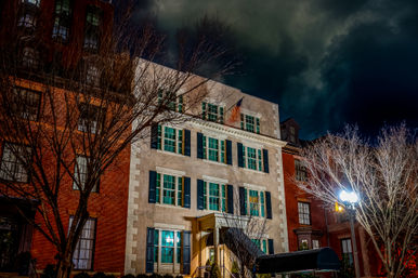 Nighttime urban scene of a historic downtown townhouse with green shutters and lit windows, bare trees and a glowing streetlamp beneath a dramatic cloudy sky.