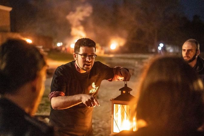 Lantern-lit storyteller addressing a small group at a nighttime outdoor bonfire event, warm orange glow from the lantern and distant fires illuminating the scene.