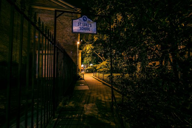 Nighttime urban school entrance with a dimly lit sidewalk flanked by a tall iron fence and dense trees, a hanging school sign overhead and warm streetlamp glow ahead.